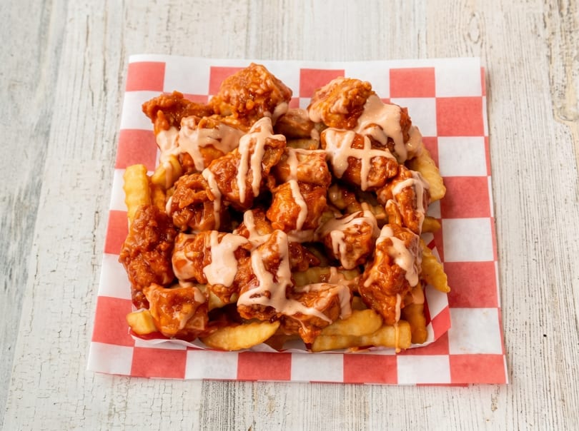 Crispy french fries topped with saucy boneless chicken wings and a creamy drizzle on checkered paper.