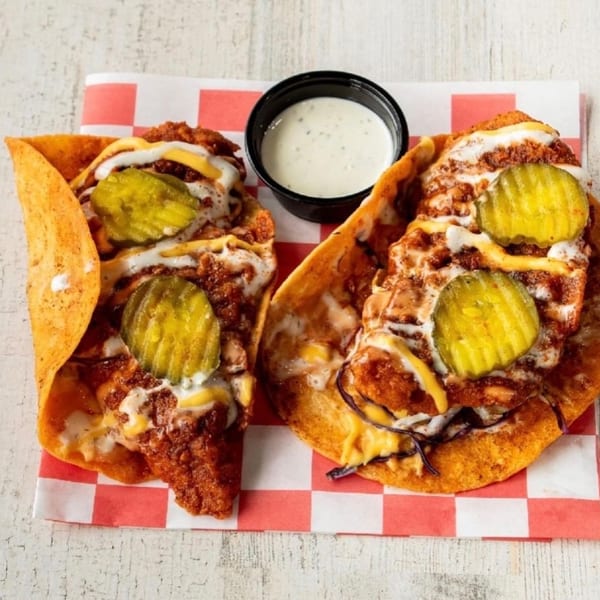 Two crispy chicken tacos with pickles, cheese, and sauce, served with a side of ranch.