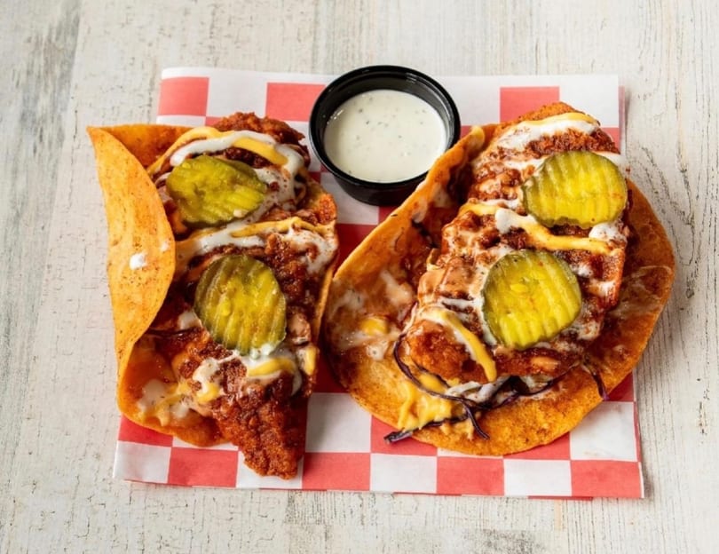 Two crispy chicken tacos with pickles, cheese, and sauce, served with a side of ranch.