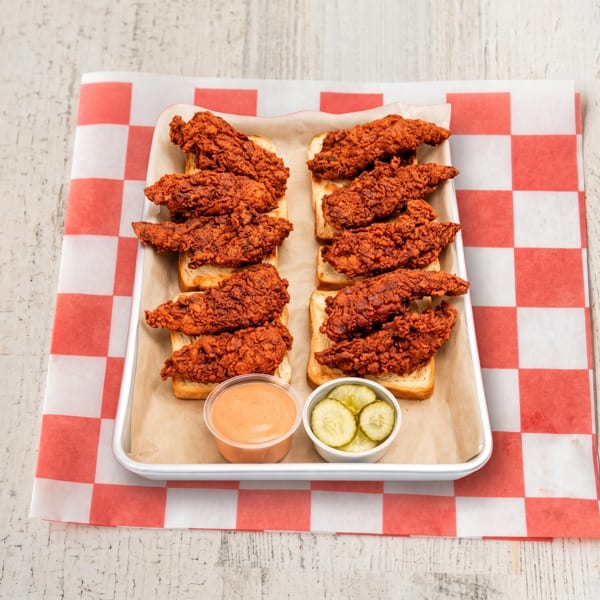 Spicy fried chicken tenders on white bread with sauce and pickles on a metal tray.