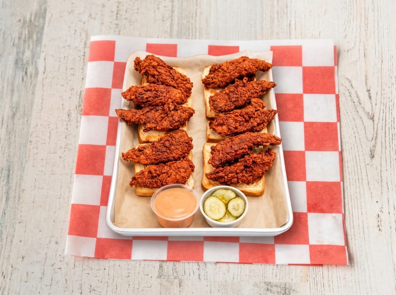 Spicy fried chicken tenders on white bread with sauce and pickles on a metal tray.